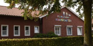 Ein Backsteingebäude mit einem roten Ziegeldach, über fünf Fenstern steht in bunten Buchstaben „St. Marien Kindergarten“, teilweise verdeckt durch eine Hecke und Äste.