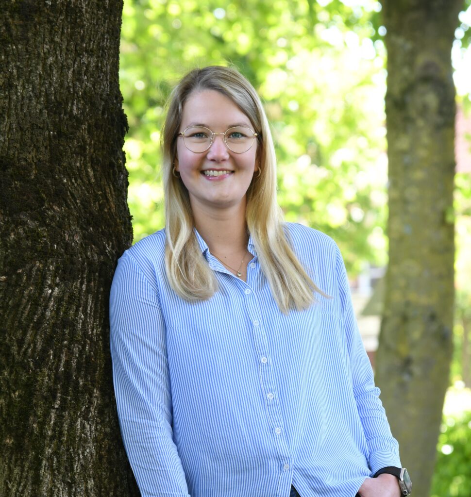 Eine junge Frau mit glatten blonden Haaren lächelt, während sie im Freien steht und sich an einen Baum mit rauer Rinde lehnt. Sie trägt eine Brille und eine hellblaue Bluse.