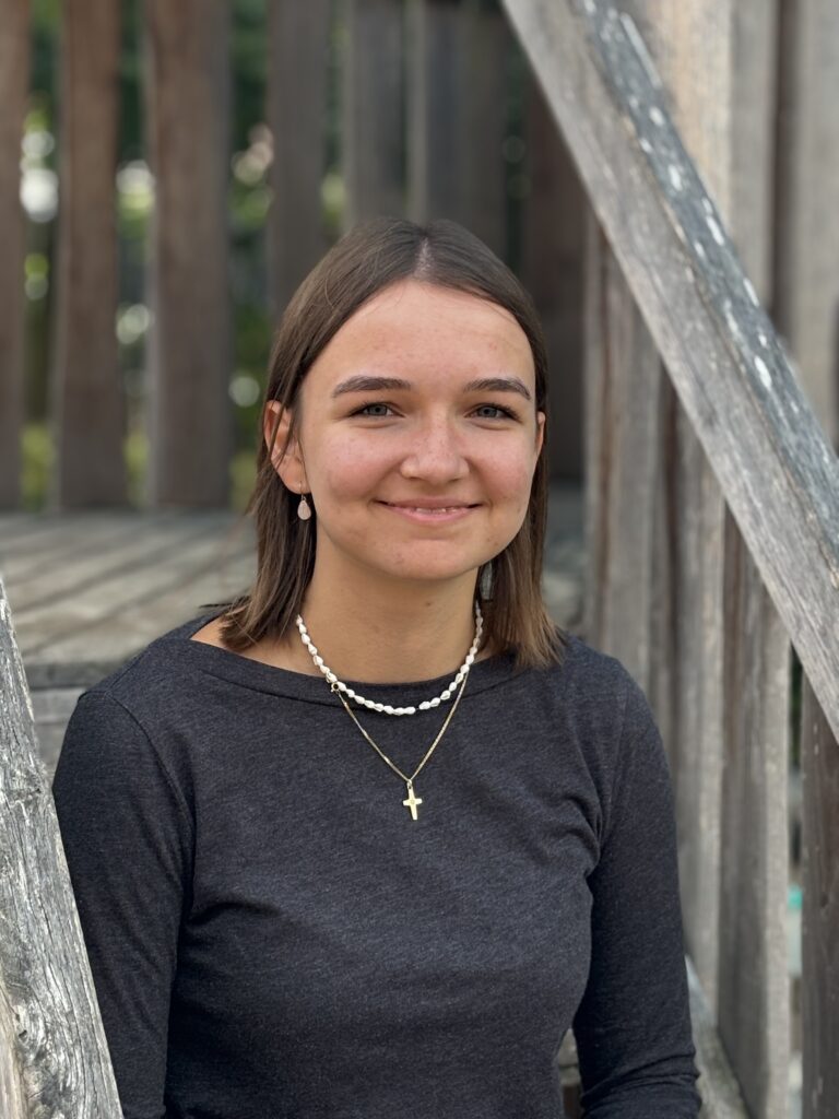 Eine junge Frau mit glatten braunen Haaren, die ein schwarzes Hemd trägt, sitz auf einer Treppe eines Spielgerüstes. Sie lächelt freundlich und schaut in die Kamera.